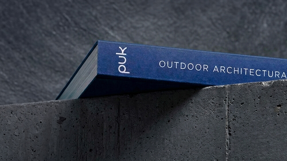 Un libro blu intitolato OUTDOOR ARCHITECTURE, con la scritta PUK sul dorso, è appoggiato su una superficie di cemento ruvida su uno sfondo grigio scuro.