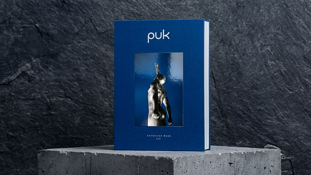 Un libro con copertina rigida blu intitolato puk è appoggiato in posizione verticale su una superficie di cemento; sulla copertina è raffigurata una foto riflettente di una statua in bianco e nero, mentre sotto l'immagine compare il sottotitolo Collection Book 2018. Lo sfondo è in pietra scura con texture.
