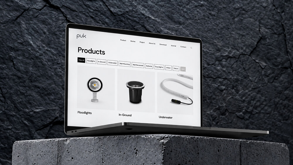 Un computer portatile è appoggiato su una superficie in pietra e mostra una pagina web dedicata ai prodotti, con immagini e categorie relative a prodotti di illuminazione quali proiettori, luci da incasso nel terreno e luci subacquee. Lo sfondo è in pietra con texture.