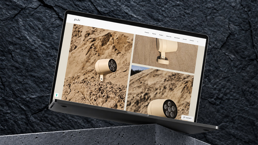 Un computer portatile appoggiato su una superficie di cemento mostra un sito web che presenta delle luci da esterno cilindriche di colore beige posizionate sulla sabbia, riprese da diverse angolazioni.