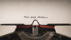 Un primo piano di una macchina da scrivere che digita “Racconta la tua storia...” su un foglio bianco, con le barre dei caratteri visibili e un nastro di inchiostro rosso e nero inserito nella macchina.