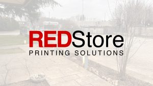 Logo per REDStore Printing Solutions con il ROSSO in grassetto e il resto in nero, sovrapposto a una foto di un'area esterna pavimentata con alberi e cielo coperto.