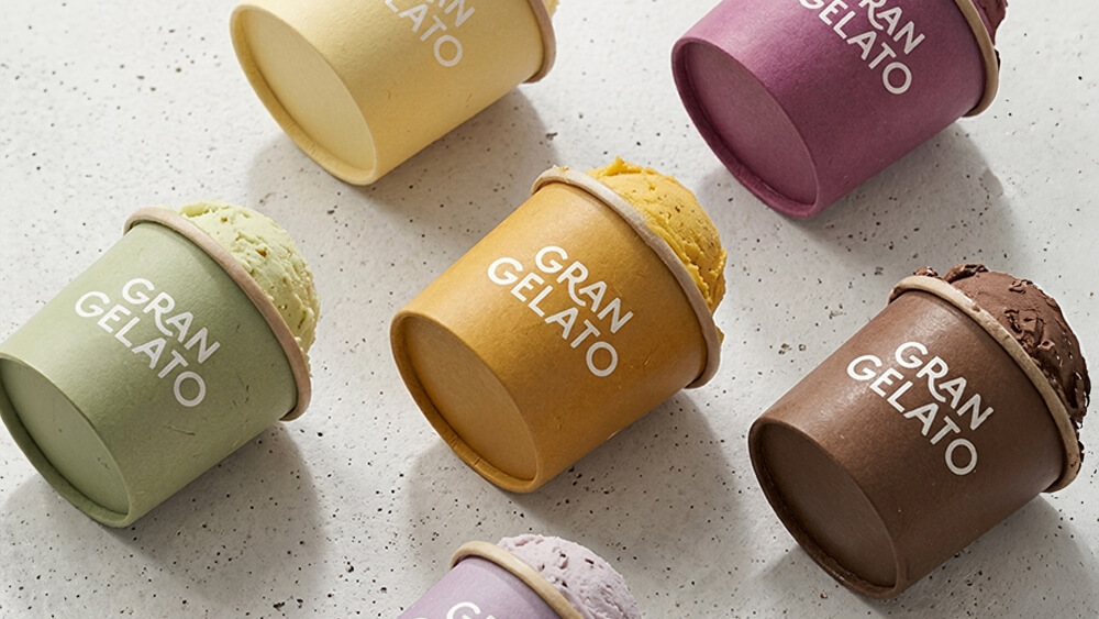 Sei coppette di gelato colorate con l'etichetta «GRAN GELATO» sono disposte su una superficie chiara maculata; ogni coppetta presenta un gusto e un colore diversi, tra cui verde, giallo, viola, marrone e crema.